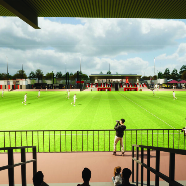Short version: Architectural visualisation of a proposed football stadium, viewed from an elevated position in a covered stand. Players in white kits are shown on a bright green pitch, with a red and white seated main stand visible on the far side.